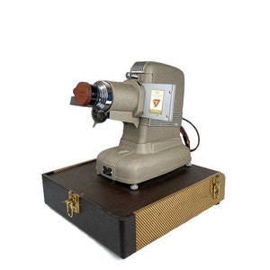 May include: A vintage silver slide projector with a brown lens cover, sitting on top of a brown and gold patterned carrying case. The projector has a label that reads "The  Illustrated  " with a triangle symbol.