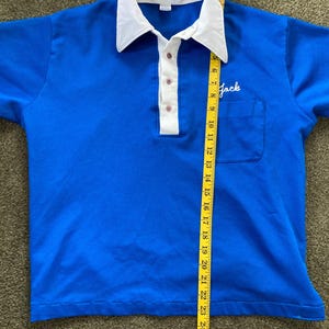 May include: A blue short-sleeved polo shirt with a white collar and a white button placket. The shirt has a small pocket on the left chest with the word "Jack" embroidered in white. The shirt is being measured with a yellow tape measure.