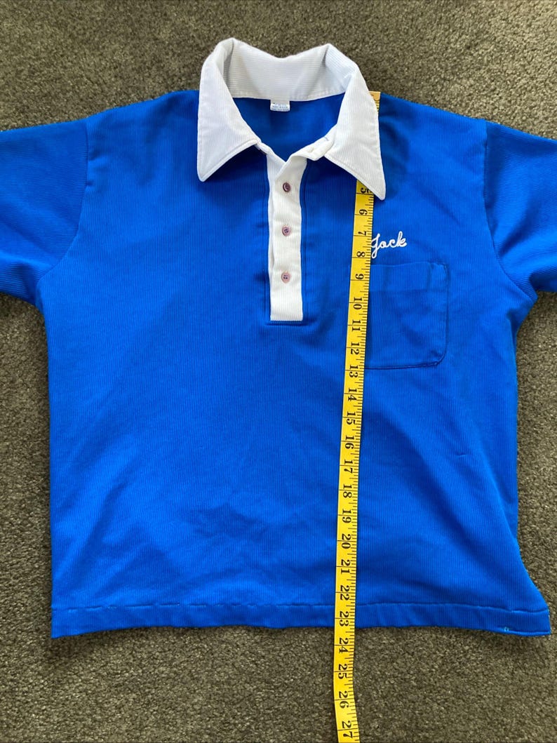 May include: A blue short-sleeved polo shirt with a white collar and a white button placket. The shirt has a small pocket on the left chest with the word "Jack" embroidered in white. The shirt is being measured with a yellow tape measure.