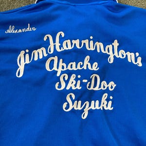 May include: A blue polo shirt with white embroidered text on the back. The text reads "Jim Harrington's Apache Ski-Doo Suzuki" and "Alexander" is embroidered on the top left of the shirt.