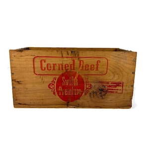 May include: A vintage wooden crate with the words "Corned Beef" in red lettering. Below, a red circular emblem reads "Swift's Premium". The crate is made of light-colored wood with visible grain and metal fasteners.
