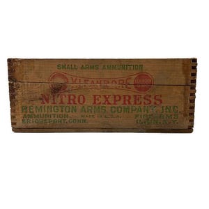 May include: A vintage wooden ammunition box with the text "NITRO EXPRESS" in red and "REMINGTON ARMS COMPANY, INC." in green. The box is made of aged wood and features the words "SMALL ARMS AMMUNITION".