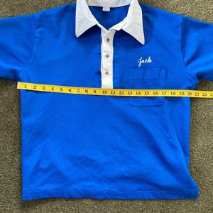 May include: A blue short-sleeved polo shirt with a white collar and a white button placket. The shirt has a pocket on the left chest with the word "Jack" embroidered in white. The shirt is measured with a tape measure, showing a length of 56 cm.