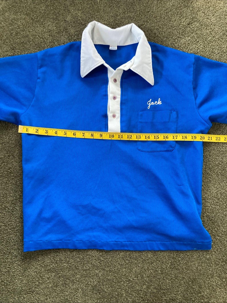 May include: A blue short-sleeved polo shirt with a white collar and a white button placket. The shirt has a pocket on the left chest with the word "Jack" embroidered in white. The shirt is measured with a tape measure, showing a length of 56 cm.