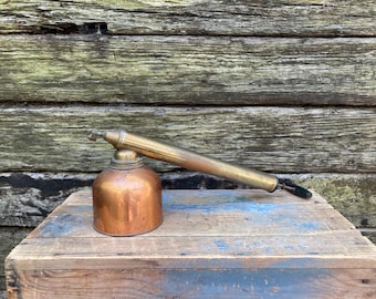 May include: An antique copper and brass sprayer with a long nozzle, resting on a wooden crate. The sprayer has a rounded copper base and a brass nozzle. The wood background has a weathered appearance.