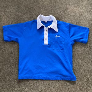 May include: A blue short-sleeved polo shirt with a white collar and a white pocket. The shirt has a button-up closure and the word "Jack" embroidered on the pocket.