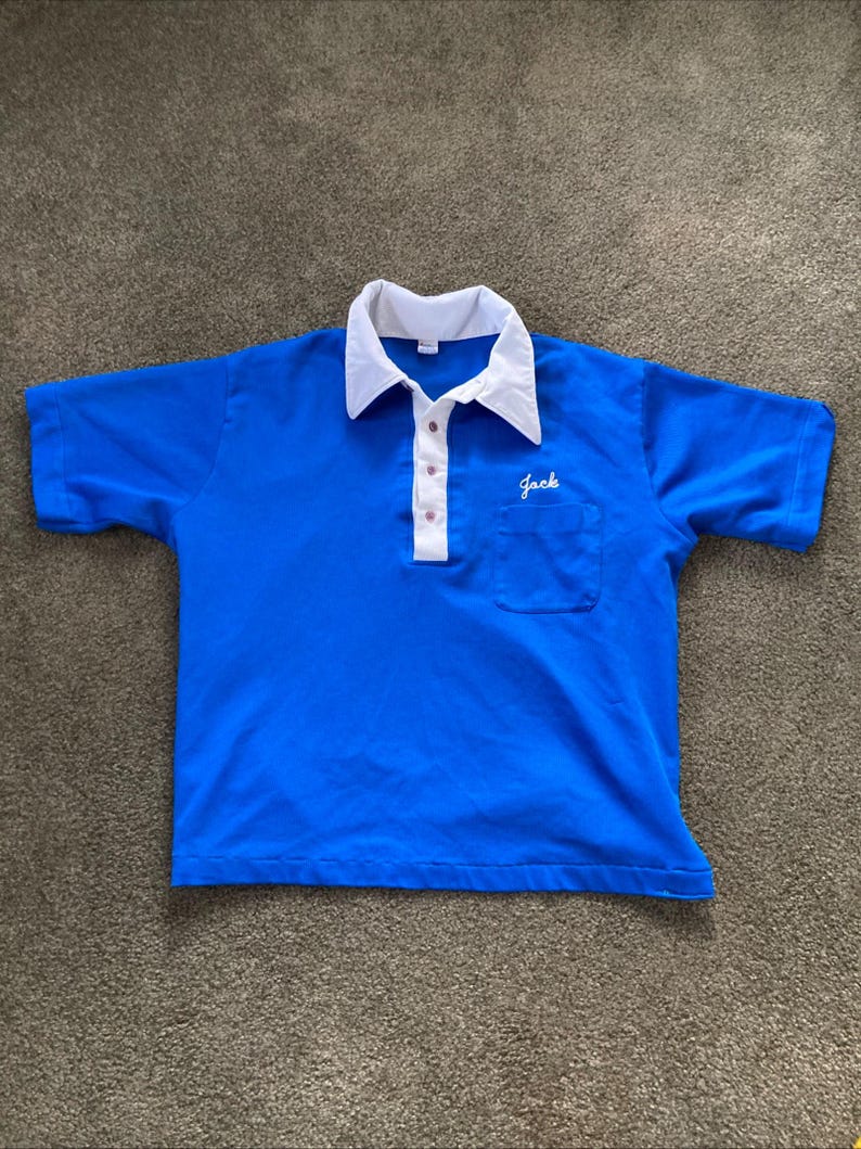 May include: A blue short-sleeved polo shirt with a white collar and a white pocket. The shirt has a button-up closure and the word "Jack" embroidered on the pocket.