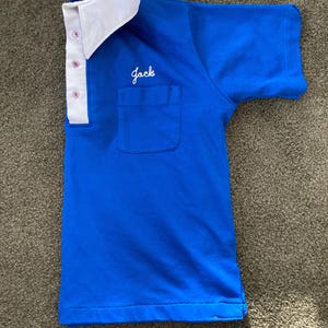 May include: A blue short-sleeved shirt with a white collar and a pocket on the left side. The shirt has a white embroidered name "Jack" on the pocket.