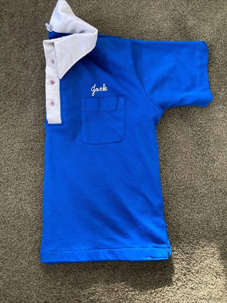 May include: A blue short-sleeved shirt with a white collar and a pocket on the left side. The shirt has a white embroidered name "Jack" on the pocket.