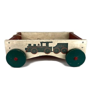 May include: A vintage wooden toy wagon with a painted train design. The wagon has green wheels with red centers, and a cream-colored body with a dark green train illustration. The wagon's interior is red.