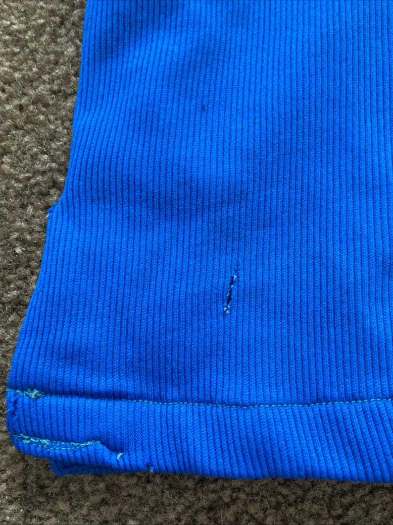 May include: A close-up of a bright blue ribbed fabric with a small hole in the centre. The fabric has a subtle sheen and a slightly textured surface.
