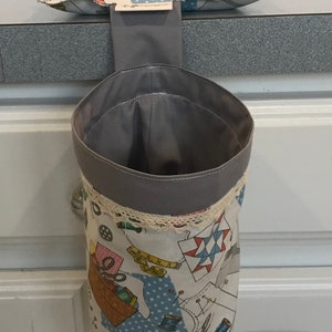May include: A gray fabric hanging storage bin with a white and blue fabric top featuring a sewing theme. The bin has a white lace trim and is hanging from a gray fabric loop.