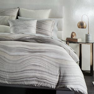 May include: A bed with a neutral-toned bedding set featuring a wavy, marbled pattern in shades of gray and white. Several pillows with textured and striped designs are arranged on the bed. A gold-toned lamp and side table are visible.