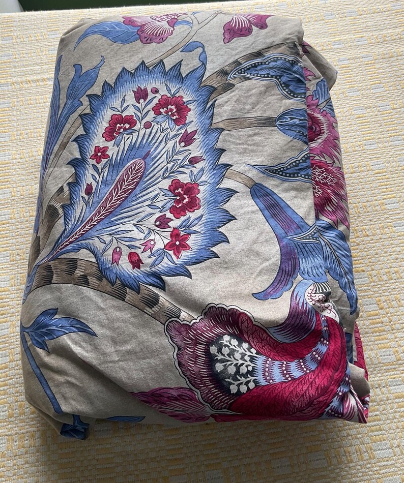 Puede incluir: Ropa de cama doblada con un estampado floral en tonos azules, rojos y morados sobre un fondo beige. El dise&ntilde;o presenta grandes flores y follaje estilizados.