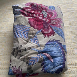 Puede incluir: Juego de cama plegado con un gran dise&ntilde;o floral. El patr&oacute;n presenta una prominente flor magenta con detalles intrincados, complementada con hojas azules y grises sobre un fondo neutro. La tela parece suave, probablemente de algod&oacute;n.