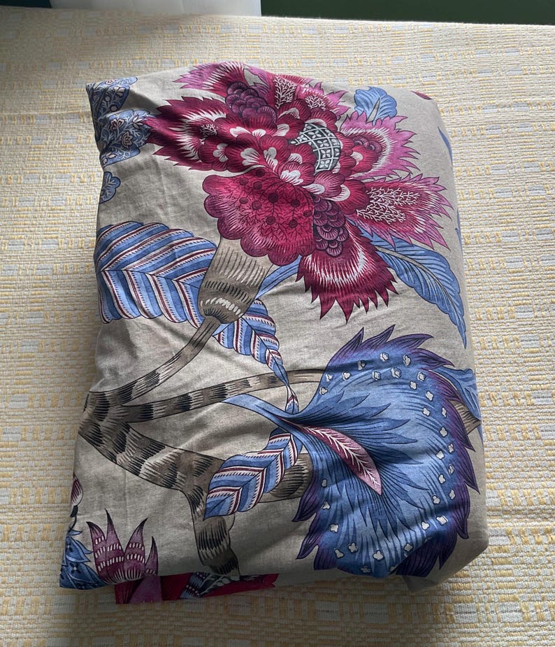 Puede incluir: Juego de cama plegado con un gran dise&ntilde;o floral. El patr&oacute;n presenta una prominente flor magenta con detalles intrincados, complementada con hojas azules y grises sobre un fondo neutro. La tela parece suave, probablemente de algod&oacute;n.