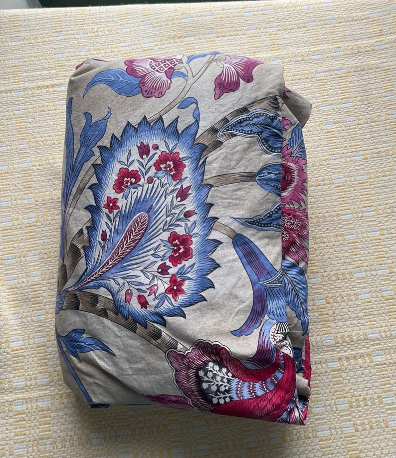 Puede incluir: Tela doblada con un estampado floral en tonos azules, rosas y grises. El dise&ntilde;o presenta grandes flores y hojas estilizadas. La tela est&aacute; doblada cuidadosamente, mostrando el intrincado patr&oacute;n.