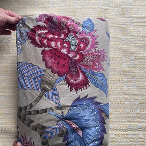 Puede incluir: Tela doblada con un gran dise&ntilde;o floral en tonos burdeos, rosa y azul sobre un fondo beige. El dise&ntilde;o incluye flores y hojas estilizadas, creando un tema bot&aacute;nico. La tela est&aacute; sujeta por una mano.