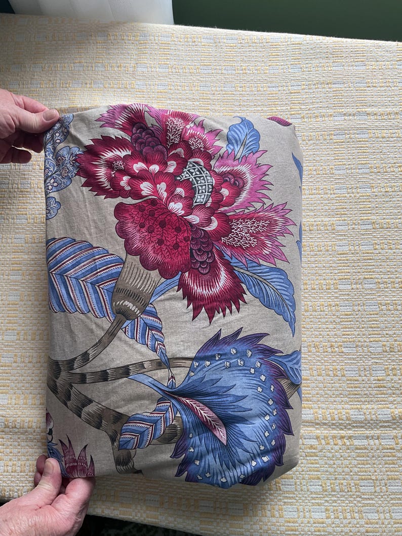 Puede incluir: Tela doblada con un gran dise&ntilde;o floral en tonos burdeos, rosa y azul sobre un fondo beige. El dise&ntilde;o incluye flores y hojas estilizadas, creando un tema bot&aacute;nico. La tela est&aacute; sujeta por una mano.