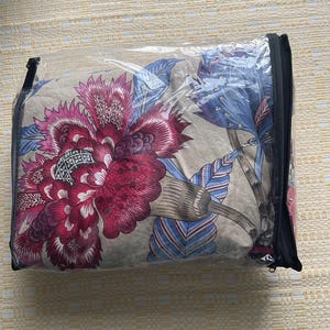 Puede incluir: Un juego de edred&oacute;n con estampado floral en una funda de pl&aacute;stico transparente con cremallera. El edred&oacute;n presenta grandes flores estilizadas en tonos burdeos, rosa y azul sobre un fondo neutro. La funda tiene una cremallera negra.