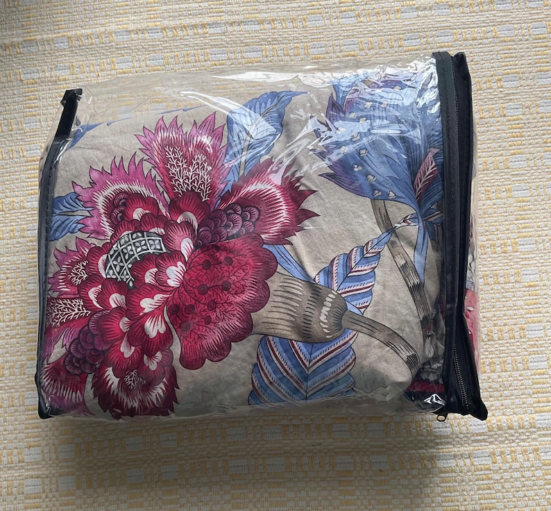 Puede incluir: Un juego de edred&oacute;n con estampado floral en una funda de pl&aacute;stico transparente con cremallera. El edred&oacute;n presenta grandes flores estilizadas en tonos burdeos, rosa y azul sobre un fondo neutro. La funda tiene una cremallera negra.