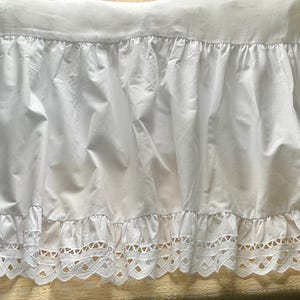 May include: A white bed skirt featuring a gathered fabric panel and a bottom border with intricate cutwork embroidery. The design includes a repeating geometric pattern along the hemline, providing a decorative finish to the plain white fabric.