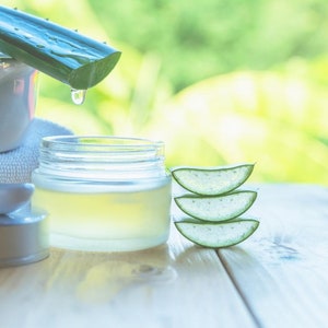 May include: A close-up of aloe vera skincare products. A clear glass jar filled with yellow liquid, aloe vera slices, and a white bowl with a spoon are on a wooden surface. A green aloe vera leaf drips water.