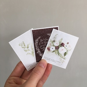 May include: Three cards with watercolor floral designs and inspirational quotes. The first card features a green fern, the second card has a burgundy background with a floral wreath and the quote "But thank be to God, which giveth us the victory." The third card has a white background with a bouquet of white and burgundy flowers and the quote "Great is the Lord, and greatly to be praised; and his greatness is unsearchable."