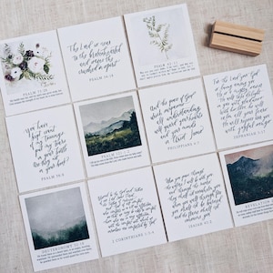 May include: A collection of square cards with inspirational quotes from the Bible, featuring calligraphy and watercolor landscape art. A wooden card holder is also present. The cards are arranged on a neutral linen surface.