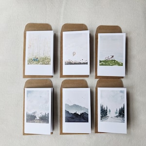 May include: Set of six blank greeting cards with brown envelopes. Each card features a watercolor illustration of a different landscape scene, including a forest, a hot air balloon, a bicycle, mountains, and a road.