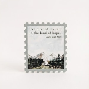 May include: A gray stamp-shaped sticker with a mountain and forest scene. The text "I've pitched my tent in the land of hope. Acts 2:26 MSG" is printed in black. The sticker has a white background.