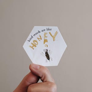 May include: A white, hexagonal sticker with the words "kind words are like HONEY" in gold lettering. A watercolor illustration of a bee is below the word "HONEY".