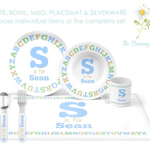 May include: A white children's dinnerware set featuring a plate, bowl, mug, placemat, spoon, and fork. Each item is decorated with the letter "S" in light blue, the name "Sean", and the alphabet in various colors.