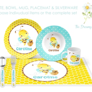 May include: A personalized children's place setting with a yellow and blue bee theme. The set includes a plate, bowl, mug, placemat, and silverware. The name "Caroline" is printed on each piece.