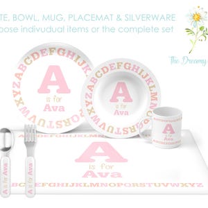 May include: A pink and gold alphabet themed children's dinnerware set with a plate, bowl, mug, and placemat. The set features the letter A and the name Ava. The Dreamy Daisy is printed on the placemat.