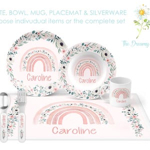 May include: A personalized children's dinnerware set with a pink and white floral design featuring a rainbow and the name "Caroline". The set includes a plate, bowl, mug, placemat, and silverware.