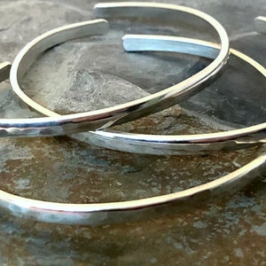 May include: Three silver hammered bangle bracelets stacked on top of each other. The bracelets are all the same width but have different diameters.