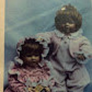 May include: Two handmade cloth dolls, one wearing a pink and white dress and holding a block, the other wearing a blue and white outfit. The dolls have brown hair and painted faces. The dolls are sitting on a white background with a set of colorful alphabet blocks.