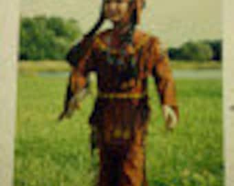 May include: A child wearing a brown and tan Native American costume with a feathered headdress. The costume includes a long-sleeved shirt, pants, and a belt with fringe. The child is holding a toy spear in their right hand.