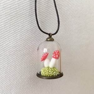 May include: A glass pendant necklace with a small, clear glass dome containing two red and white spotted mushrooms on a green moss base.