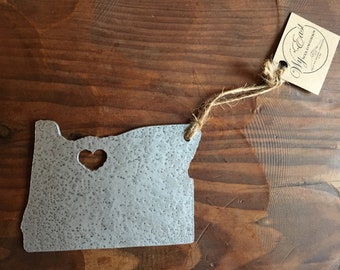 Puede incluir: Un adorno de metal plateado con forma del estado de Oregón con un corazón recortado en el centro. El adorno está colgado de una cuerda de hilo con una etiqueta de papel marrón que dice "Wy'East Welded Works".