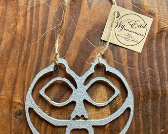 Puede incluir: Un adorno de calabaza de metal plateado con una cara tallada. El adorno está colgado de una cuerda de yute y tiene una etiqueta adjunta que dice "Wy'East Weldworks, Mount Hood, OR, USA".