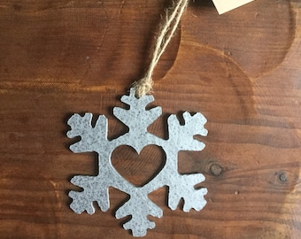 Puede incluir: Un adorno de copo de nieve de metal plateado con un corazón recortado en el centro. El adorno cuelga de una cuerda de hilo y tiene una etiqueta que dice "My East Weldworks, Mount Horeb, WI".
