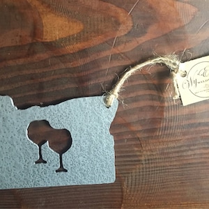 May include: A metal cutout of the state of Oregon with two wine glasses cut out inside. The cutout is attached to a brown twine string with a tag that reads "Wy'East Woodworks".