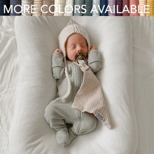 May include: A sleeping infant wearing a light green onesie and a knitted bonnet. The baby has a pacifier and a beige muslin security blanket. The text "MORE COLORS AVAILABLE" is at the top of the image.