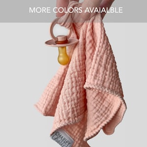 May include: A light peach-colored muslin fabric square attached to a matching peach-colored pacifier with a translucent amber-toned nipple. The fabric has a textured, crinkled appearance and a white label reading CHARLEY CHARLEY. Text reads MORE COLORS AVAIIALBLE.