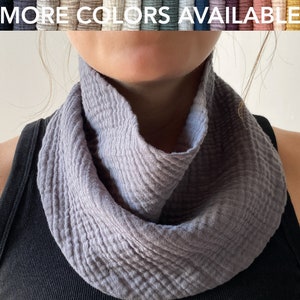 May include: A gray, textured infinity scarf made of soft fabric. The scarf is draped around the neck, with the text "MORE COLORS AVAILABLE" displayed above. The scarf appears to be a light gray color.