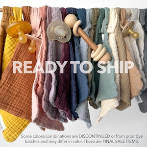 May include: A variety of colors of soft, textured fabric squares with a wooden rattle and pacifiers. The text "READY TO SHIP" is displayed in the center of the image. The text "Some colors/combinations are DISCONTINUED or from prior dye batches and may differ in color. These are FINAL SALE ITEMS." is displayed at the bottom of the image.