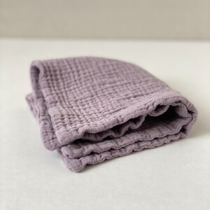 May include: A folded square of mauve-colored muslin fabric featuring a distinct crinkled, multi-layered texture. The textile displays a grid-like pattern created by the stitching of the soft, lightweight material, showing multiple layers of fabric.