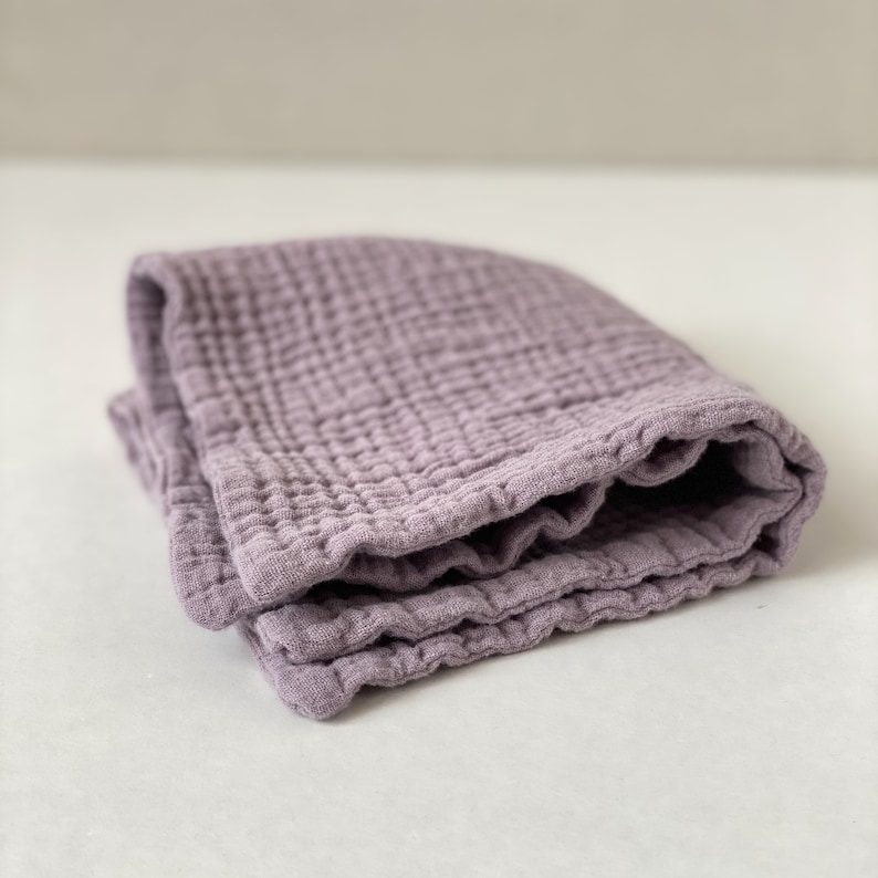 May include: A folded square of mauve-colored muslin fabric featuring a distinct crinkled, multi-layered texture. The textile displays a grid-like pattern created by the stitching of the soft, lightweight material, showing multiple layers of fabric.